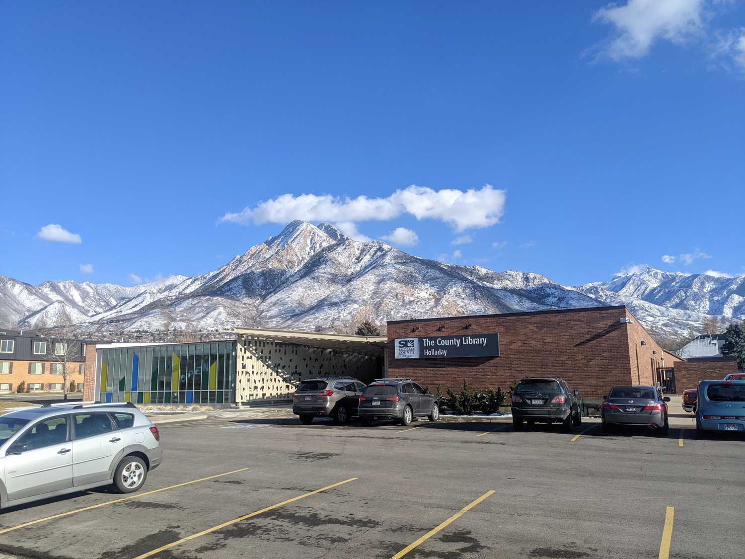 A parking lot with cars and a building with snow covered mountains in the background.