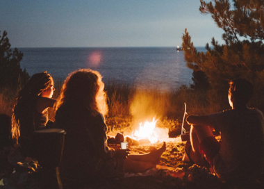 A group of people sitting around a fire by the water.