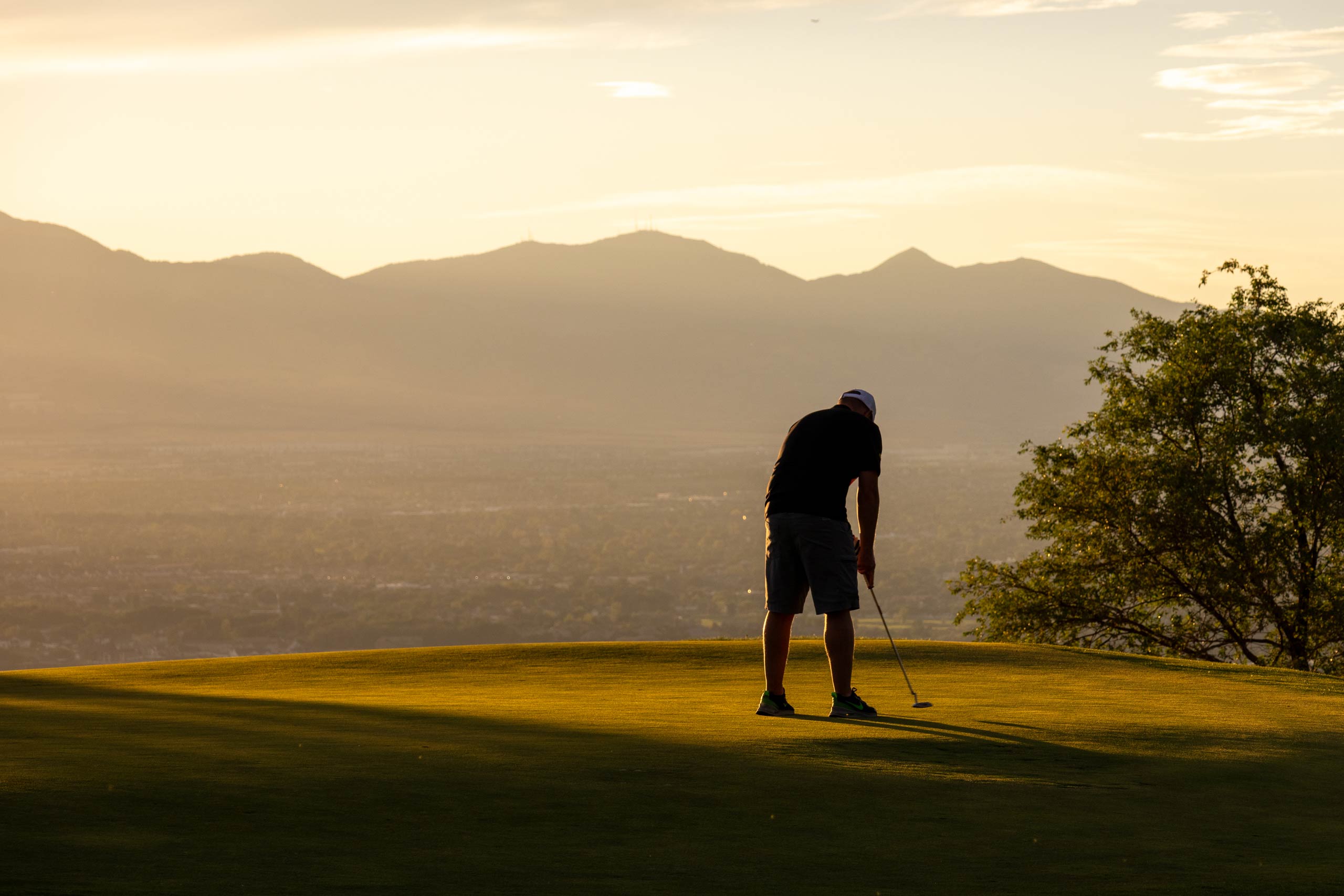 A man putting on a golf course green with mountains in the distance at sunset.