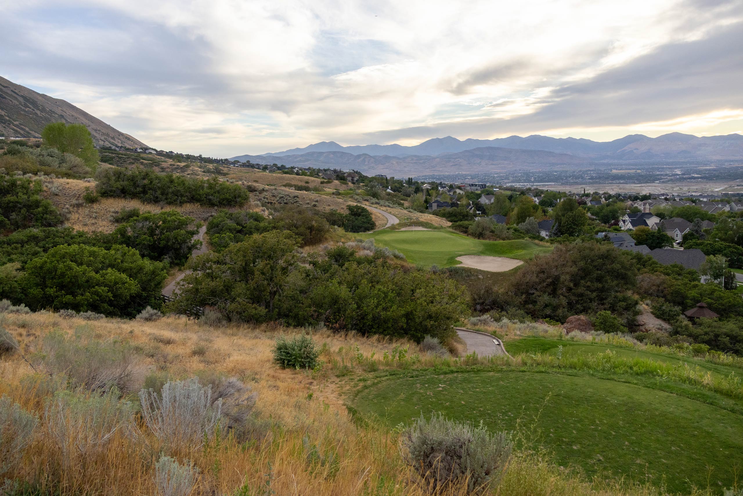 A landscape view of South Mountain golf course with trees and grass, and mountains and Draper Utah in the background.