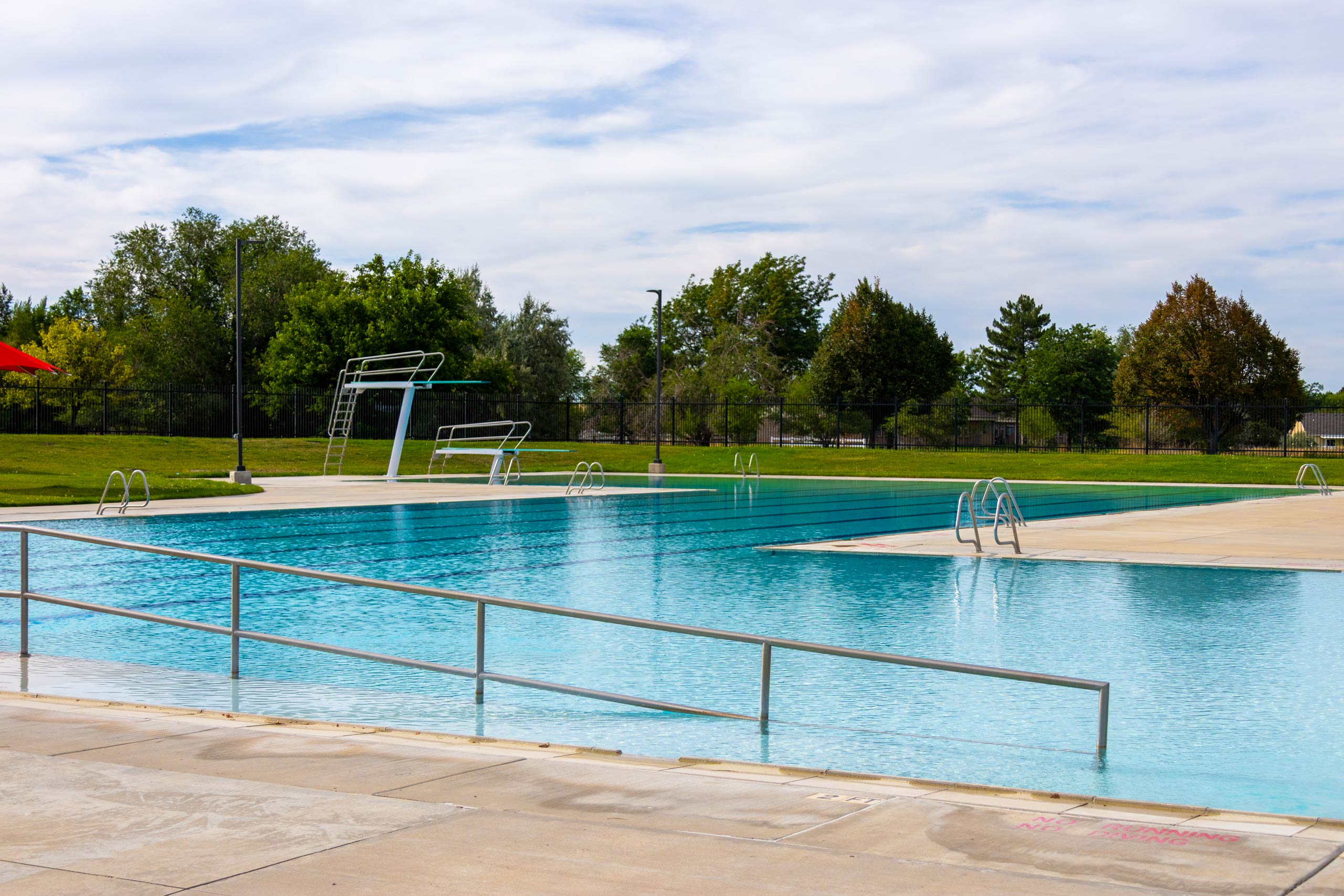 A view of diving boards, lap lanes, and ramp entrance of the South County Outdoor Pool in Riverton, Utah, with trees and grass in the background.