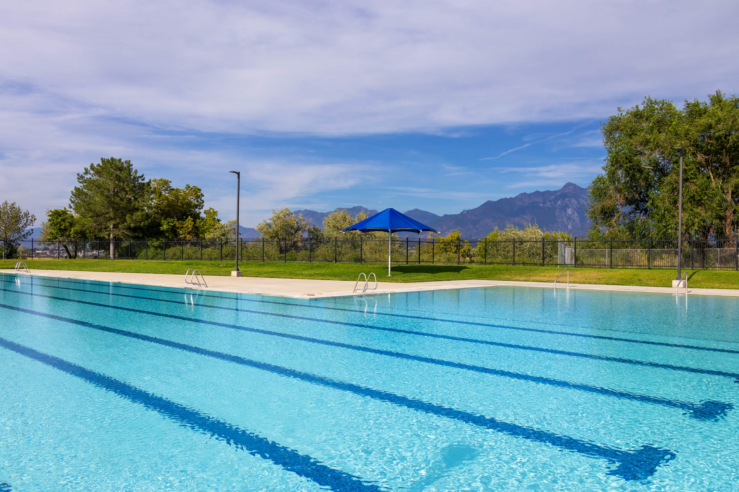 Lap lanes and relaxing area with a blue umbrella at South County Outdoor Pool in Riverton, Utah, with mountains in the distance.