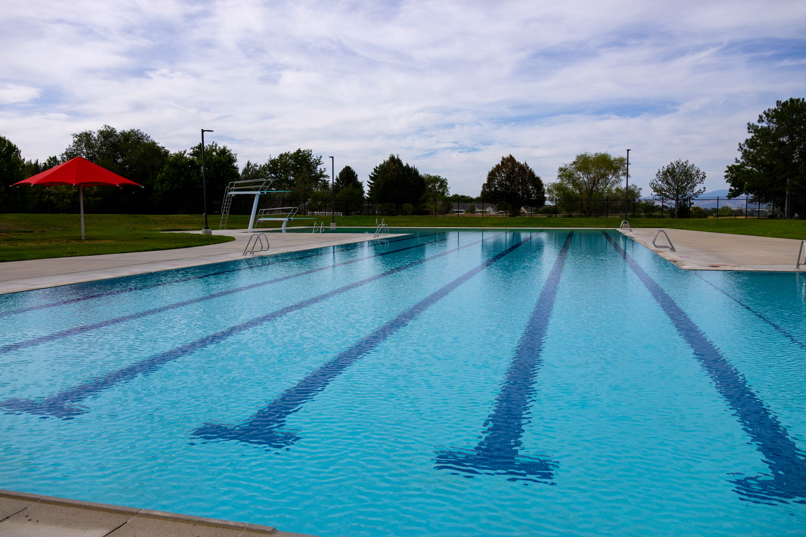 Swimming lanes, diving boards, and an umbrella at South County Outdoor Pool in Riverton, Utah, surrounded by trees.